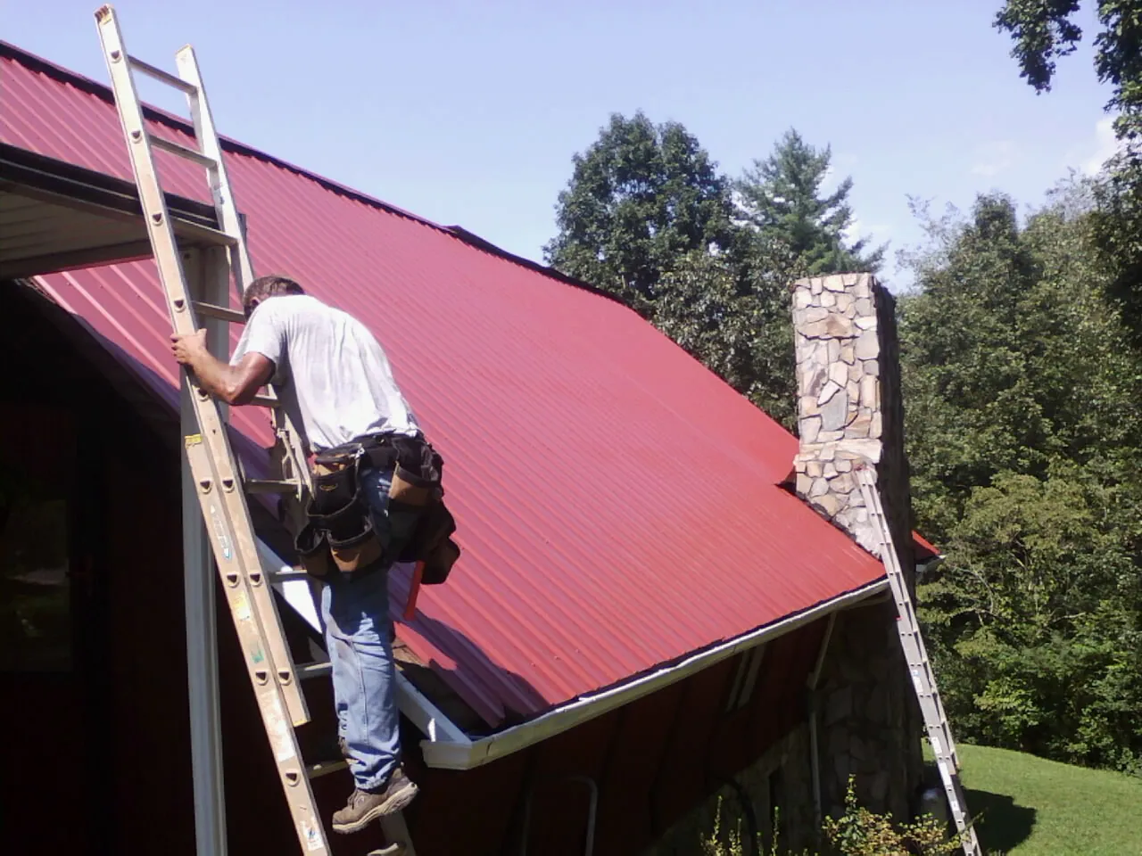 Roofer on a ladder beside a red metal roof and stone chimney — Soffit & Fascia Repair by Roofing Brothers in Elizabeth City